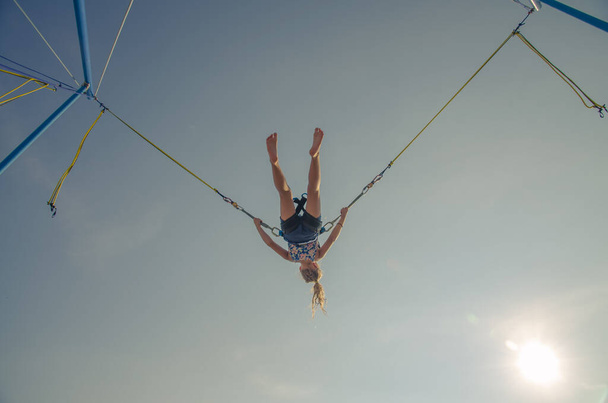 active little young teen girl jumping in trampoline high into the blue sky safed with  elastic safety belt - Photo, Image