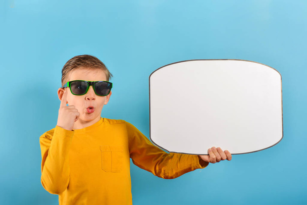 Niño sorprendido en gafas sostiene un cartel blanco con texto y levantó el dedo hasta la parte superior sobre un fondo azul
. - Foto, Imagen