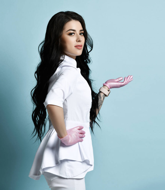 Young brunette woman cosmetologist professional in uniform is posing sideways with her hand up with open palm - Zdjęcie, obraz