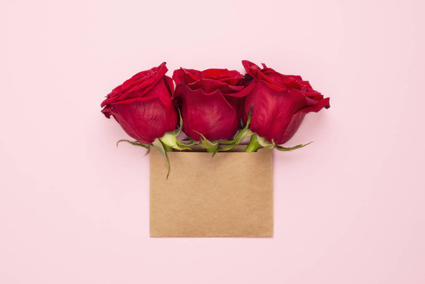 Τρία έντονα κόκκινα τριαντάφυλλα σε καφέ φάκελο με αντίγραφο χώρου. Trendy banner για την Ημέρα του Αγίου Βαλεντίνου, Παγκόσμια Ημέρα της Γυναίκας ή την ημέρα των μητέρων. Πάρτι ή πρόσκληση γάμου. Πάνω άποψη, επίπεδη lay - Φωτογραφία, εικόνα
