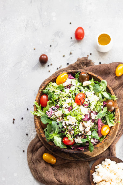 Vitamin salad with green mix salad leaves, colored cherry tomatoes, ricotta or feta cheese and balsamic in a wooden plate on a light grey background. Healthy food concept. - Photo, Image