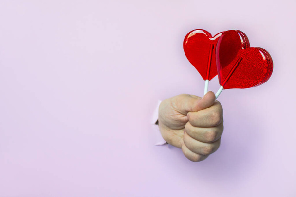 Two red lollipops on a heart shaped stick in your hand through a hole on a pink background with copy space - Fotoğraf, Görsel