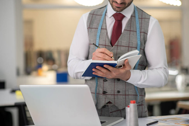 Male tailor standing near laptop, making notes - Φωτογραφία, εικόνα