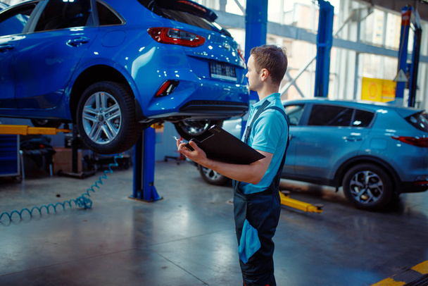 Repairman with a checklist stands at vehicle on lift, car service station. Automobile checking and inspection, professional diagnostics and repair - Photo, image