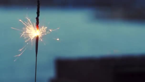 Burning, orange, flame, defective sparkler with sparks in the afternoon, morning on the street, outdoors in defocusing, on a blue background of the river, the other shore. - Video