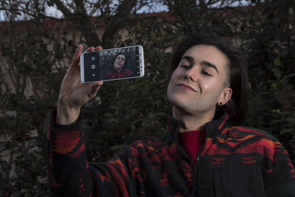 молодий чоловік фотографує свій мобільний телефон перед зеленим кущем червоних ягід
 - Фото, зображення