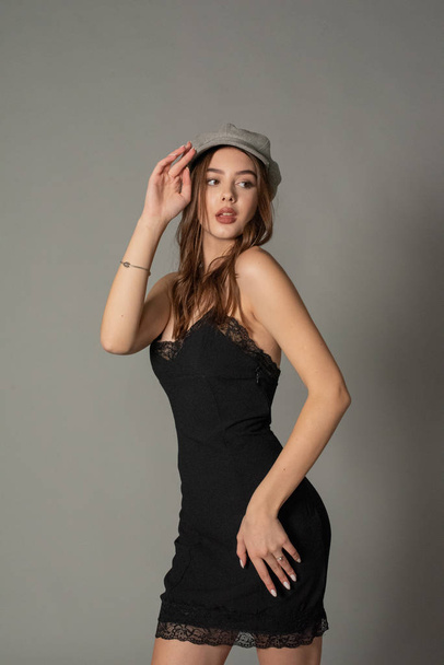Glamorous young woman wears fashionable dress posing with fluttering hair at grey wall. Girl put forward hand in a black dress and a gray cap. Beautiful young girl with brown curly hairs. portrait - Фото, зображення
