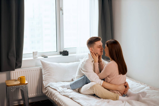 Lovely romantic man and woman going to kiss, look at each other as sit on bed - Photo, Image