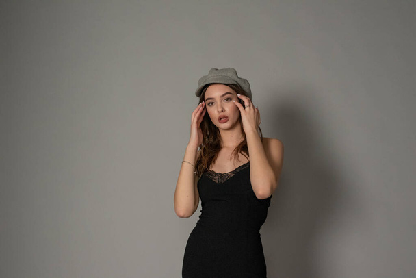 Λαμπερή νεαρή γυναίκα φοράει μοντέρνο φόρεμα που ποζάρει με φτερουγίσματα μαλλιών σε γκρι τοίχο. Το κορίτσι έβαλε μπροστά το χέρι σε ένα μαύρο φόρεμα και ένα γκρι καπέλο. Όμορφη νεαρή κοπέλα με καστανά σγουρά μαλλιά. πορτρέτο - Φωτογραφία, εικόνα