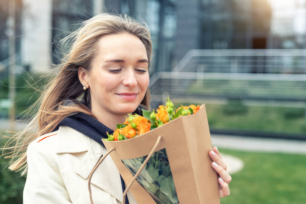 Portrait young adult smiling classy woman model hand holding authentic fresh orange spray roses flower bouquet wrapped in craft paper bag on city urban street outdoor. Spring present gift dilevery. - Photo, Image