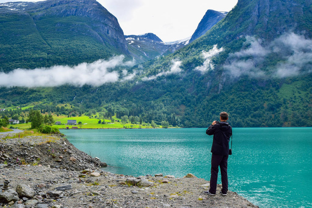 The man is taking pictures beautiful landscape of Oldevatnet glacial lake and foggy mountains in which there are glaciers of Jostedalsbreen National Park. Norway. - Fotó, kép