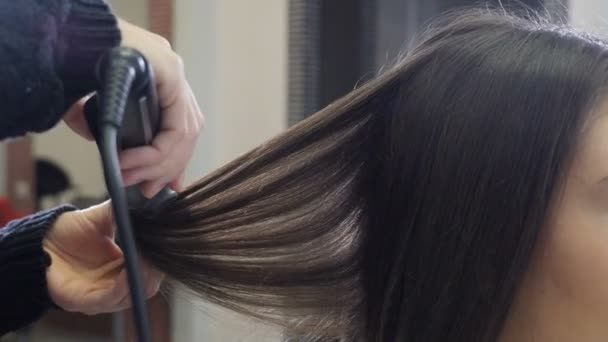 Hairstylist combing and straightening hair with hair iron in hairdressing salon. Close-up. Hairdresser doing female hairstyle with hot tongs in barbershop. Beauty and style concept. 4 k footage - Filmmaterial, Video