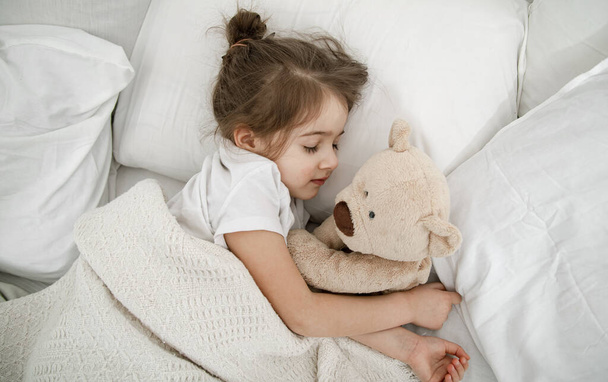 A cute little girl is sleeping in a bed with a Teddy bear toy . Concept of child development and sleep. The view from the top. - Foto, Imagem