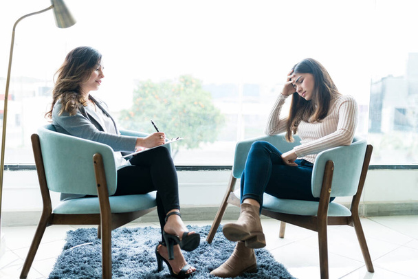 Vrouwelijke counselor schrijven op notitieblok terwijl depressieve vrouw zit met hoofd in de hand op kantoor tijdens therapiesessie - Foto, afbeelding