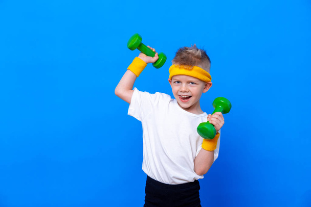 Fitness child. Portrait of sporty little boy with dumbbells over blue studio background. Gym workout. Child sportsman. Childhood activity. Sport. Fitness, health and energy. Success. - 写真・画像