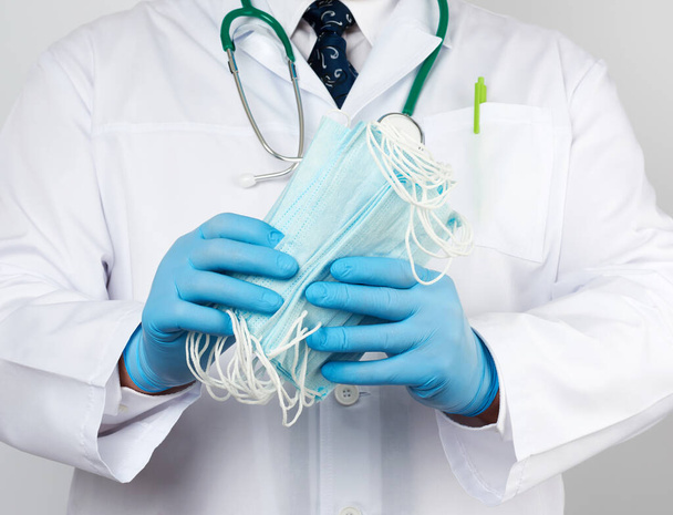 doctor in a white coat, blue latex sterile gloves holds textile medical masks in his hand, protective accessory against viruses and bacteria, close up - Fotoğraf, Görsel