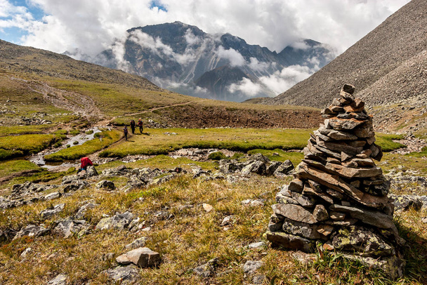 A large cairn in the foreground of a mountain landscape. Low white clouds over the mountains. Tourists with backpacks go along the path along the slope. A lot of stones on the ground. Horizontal. - Photo, image