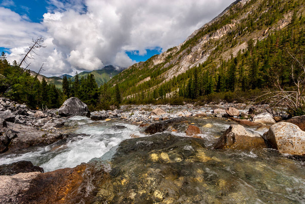 A mountain river with a wide channel flows over the stones. The river has a rocky bottom. Green forest on the hillsides and low white clouds on a blue sky. Horizontal. - Foto, imagen