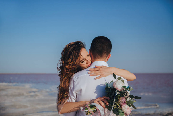 beautiful bride in a wedding dress on the seashore with a bouquet hugging her groom. Back view of the groom - Foto, Imagem