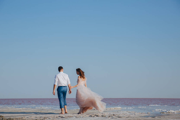 newly wed couple holding their hands walking by the sea shore. Bride in ivory flying dress. Back view - Foto, imagen