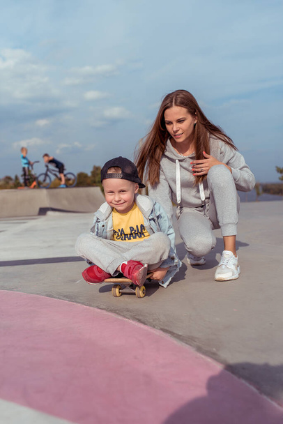 Woman mom, trains on skateboard little boy son 3-5 years old, learning to ride a skateboard, in the summer on sports ground, emotions of happiness, fun, relaxation and pleasure. Casual wear. - Foto, Imagem