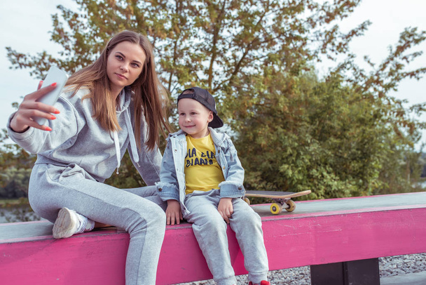 Woman mom takes pictures on phone, selfie, little boy son 3-5 years old, relax with skateboard, summer on sports ground, emotions happiness, fun, relaxation pleasure. Record video smartphone. - Foto, Bild
