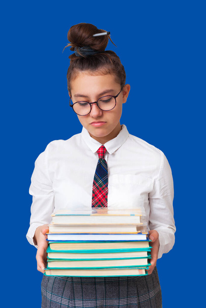 schoolgirl holds books in her hands and looks displeased. doesn't want to study. isolated on blue background - Photo, Image