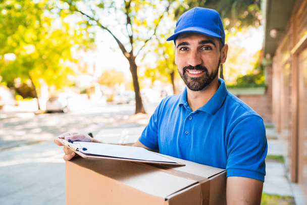 Portrait of a delivery man carrying cardboard boxes and checking list outdoors. Delivery and shipping concept. - Fotografie, Obrázek