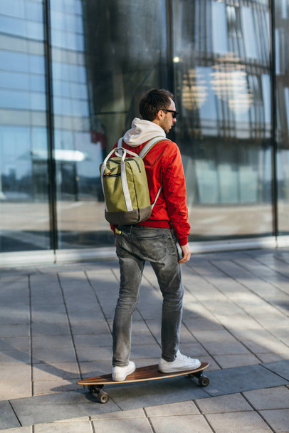 Stylish hipster in red jacket, sunglasses, jeans, white sneakers and with green backpack riding on longboard. Selective focus. Concept of leisure activity, urban. Street and lifestyle photo. - Foto, afbeelding