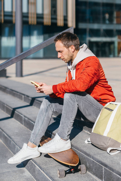 Portret van een man met baard in rood jasje, witte sneakers en groene rugzak. Hipster met skateboard zittend op treden en chatten met de mobiele telefoon. Selectieve focus. Stads- en straatfoto.  - Foto, afbeelding
