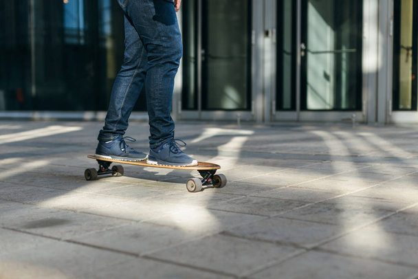 Hipster in dark blue jeans and sneakers riding on longboard in motion on asphalt. Building on the background. Selective focus on skateboard. Sunlight. Concept of leisure activity, urban and sport. - Photo, Image