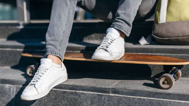 Stylish hipster in grey jeans and white sneakers put his feet on the longboard. Male resting sitting on the steps under the sun. Urban and street photo. Healthy lifestyle concept. - Photo, Image