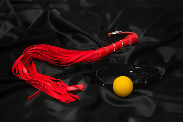 Látigo rojo y mordaza sobre un fondo de seda negro. Accesorios para juegos sexuales para adultos
. - Foto, imagen