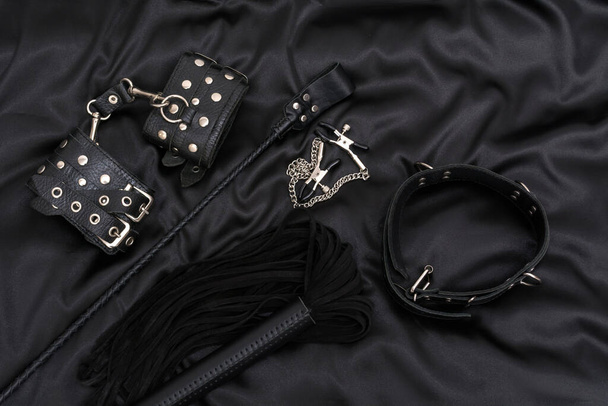 Leather handcuffs, black whip, chain collar, nipple clamps and stack on black background. BDSM outfit. - Photo, image