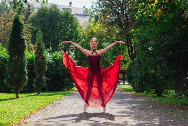 Woman ballerina in red ballet dress dancing in pointe shoes in autumn park. Ballerina standing in beautiful ballet pose - 写真・画像