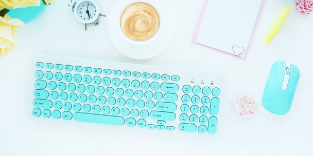 The computer keyboard and mouse are white and turquoise, a clipboard, a cup of coffee, an alarm clock and flowers. Home office concept. Banner, flat layout, top view - Photo, Image