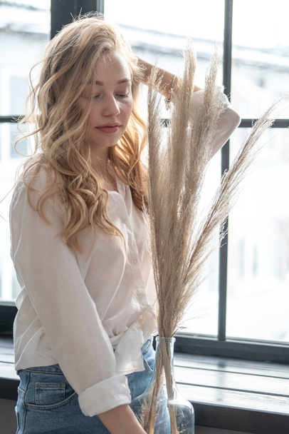 Young beutiful blond woman wearing white shirt and blue jeans standing next to window. Morning portrait of a girl holding vase with dry plants. - Foto, Bild