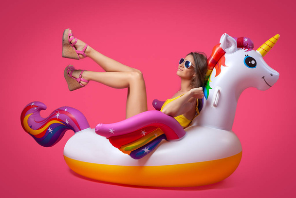Beautiful slim woman in a summer clothes, having fun and enjoying life on a white inflatable unicorn on a pink isolated background. Concept summer mood, relaxation and beauty. - Photo, Image