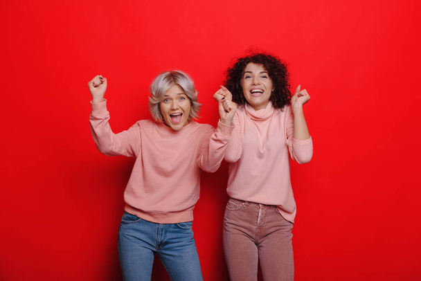 Curly cabelos caucasianos menina está se sentindo feliz perto de sua amiga loira enquanto posando em um fundo vermelho
 - Foto, Imagem