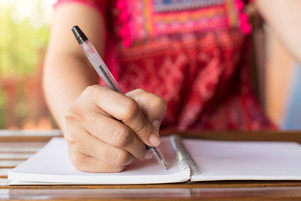 Una mujer sosteniendo un bolígrafo está escribiendo en un cuaderno y una taza de café en una mesa de madera con luz cálida
 - Foto, Imagen