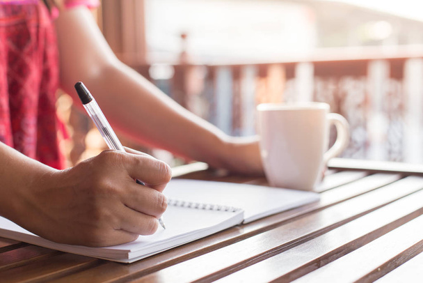 A woman holding a pen is writing on a notebook and a coffee mug on a wooden table with warm light - Photo, Image