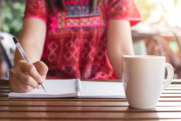 Eine Frau mit Stift schreibt auf einem Notizbuch und einem Kaffeebecher auf einem Holztisch mit warmem Licht. - Foto, Bild