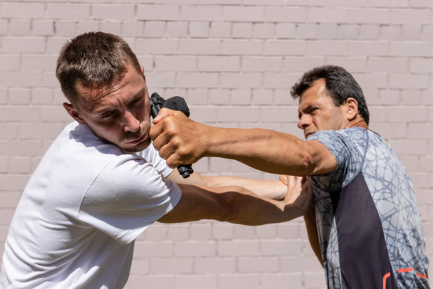 Martial arts instructors Krav Maga demonstrate self-defense techniques in a street fight. Defender hits an attacker in the face with a gun taken from him, close-up - Foto, Imagem