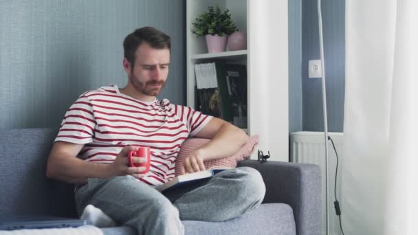 Junger Mann sitzt auf Sofa im Wohnzimmer und liest Buch mit Tee oder Kaffeetasse - Filmmaterial, Video