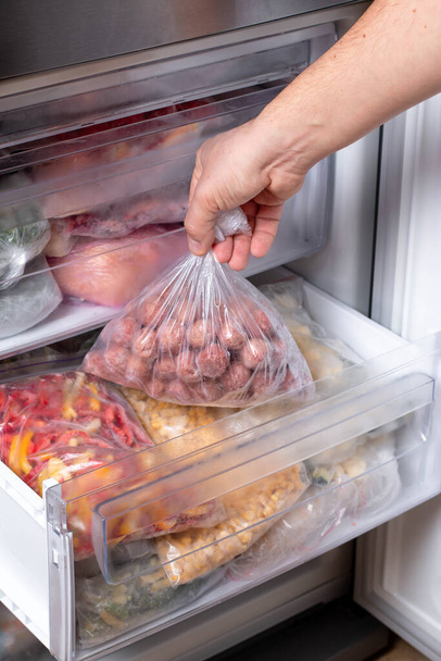 Man putting plastic bag with meatballs in refrigerator with frozen vegetables, closeup - Photo, Image
