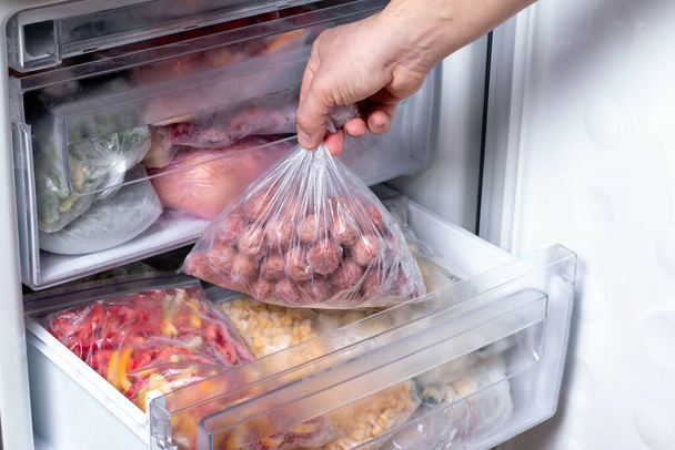 Man putting plastic bag with meatballs in refrigerator with frozen vegetables, closeup - Photo, Image