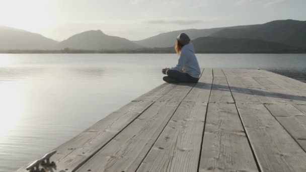 Μια σκεπτική νεαρή γυναίκα με μακριά μαλλιά που φαίνεται από το πλάι κοιτάζει τη λίμνη το ηλιοβασίλεμα καθισμένη σε μια παλιά ξύλινη προβλήτα. - Πλάνα, βίντεο