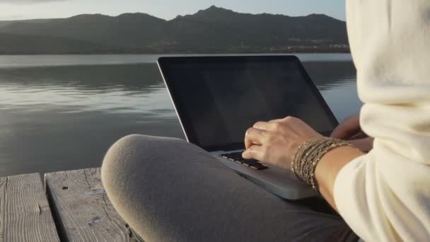 Koncepcja wyboru pracy przez internet: młoda kobieta pracuje na laptopie siedząc na molo nad jeziorem o zachodzie słońca - Materiał filmowy, wideo