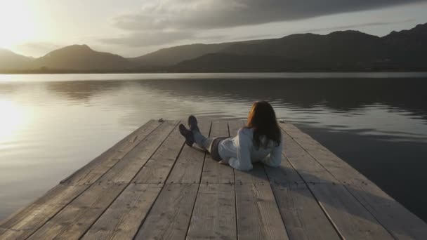 Zamyślona młoda kobieta z długimi włosami widziana od tyłu patrzy na jezioro o zachodzie słońca leżące na starym drewnianym molo - Materiał filmowy, wideo