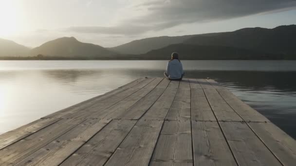後ろから見た長い髪の若い女性は、古い木製の桟橋に座って日没で湖を見て - 映像、動画